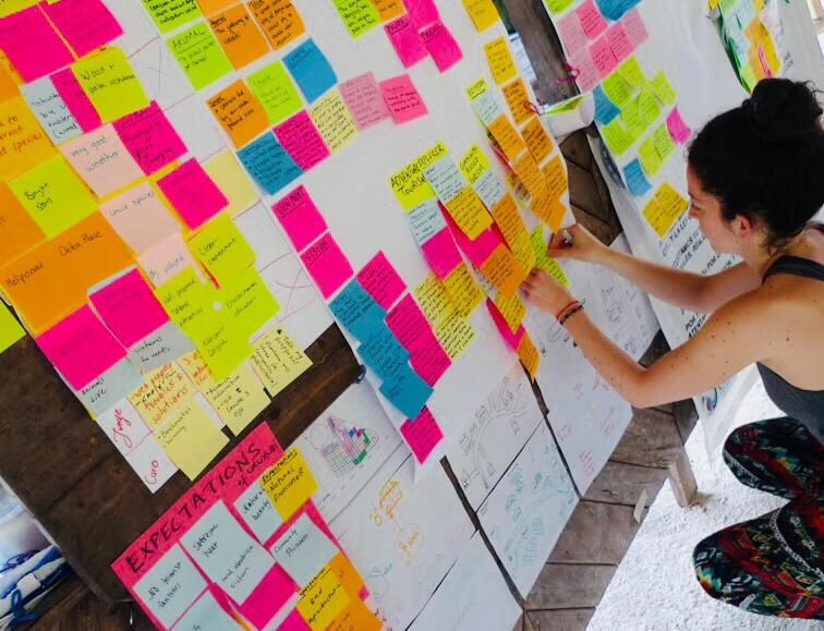 A white woman with dark hair wearing a grey tank top and patterned trousers places brightly colored sticky notes on a board. The sticky notes have handwritten text on them.