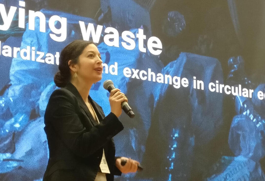 A white woman with dark hair wearing a black blazer and grey skirt is standing on stage holding a microphone on the left side of the photo. There is a blue background with pictures of trash and text that says "Buying waste: Standardization and exchange in